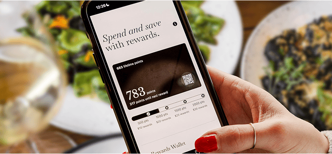A hand holds a smartphone displaying a rewards app with 783 points available, in a restaurant setting with food and a glass of wine on the table.