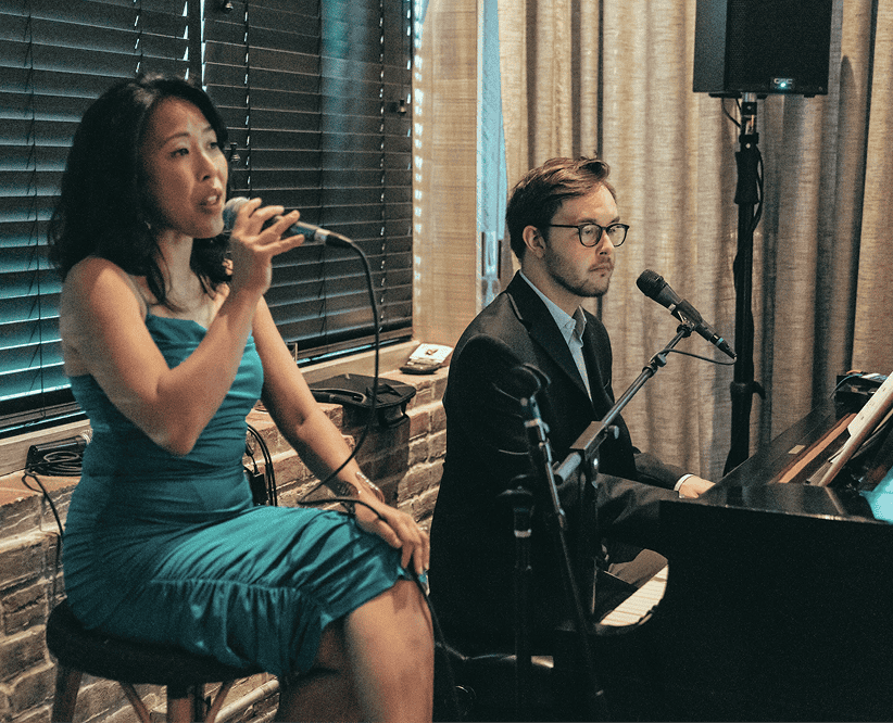 A woman in a teal dress sings into a microphone while a man in a suit plays piano beside her in an indoor setting.
