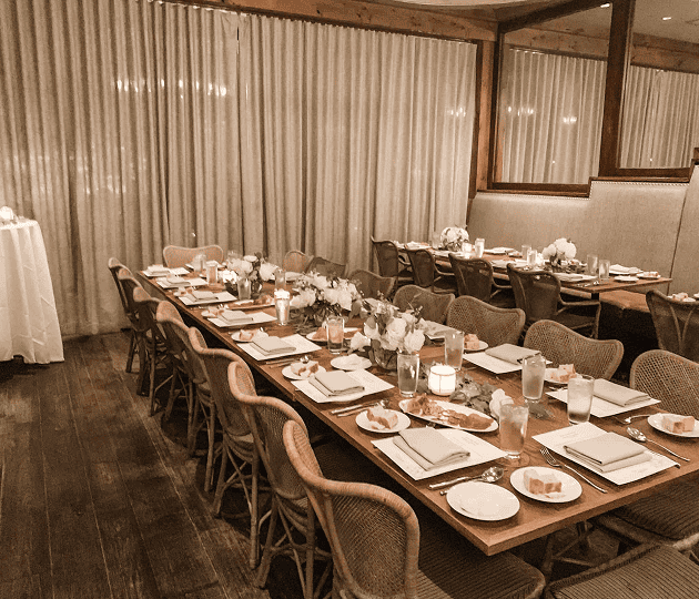 A long dining table is set with plates, glasses, napkins, candles, and white floral centerpieces in an elegant, softly lit restaurant room.