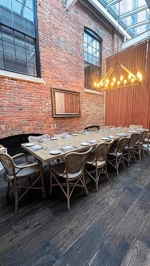A long wooden dining table with ten chairs is set with plates and glassware in a room with exposed brick walls and large windows.
